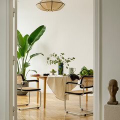 Obraz premium Inviting dining area with natural light, modern chairs, round table, and abundant green plants for a fresh, cozy atmosphere