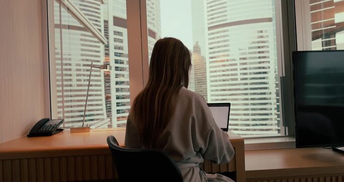 Woman typing on laptop at desk in modern office overlooking urban skyscrapers during daytime. Back view shot showing city skyline through large window. Professional remote work environment.