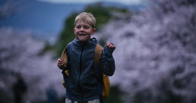 Happy little boy putting on backpack in front of spring cherry blossom trees - slow motion medium shot