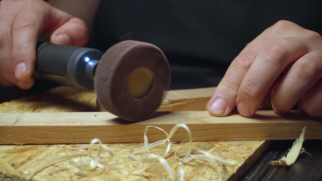 Craftsman uses rotary tool on wooden plank, creating shavings while shaping the wood in a workshop with tools and materials visible in the background