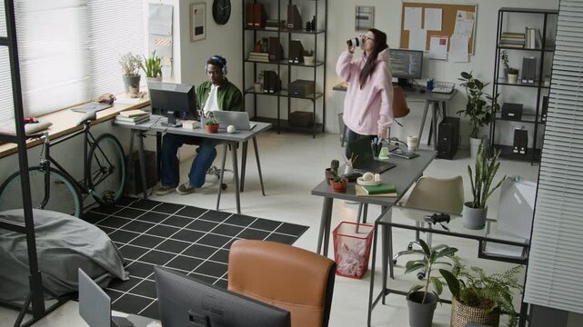 Wide overhead surveillance shot of diverse software developers working on computers at technology startup office, Caucasian woman walking for coffee, interacting with black male colleague on return