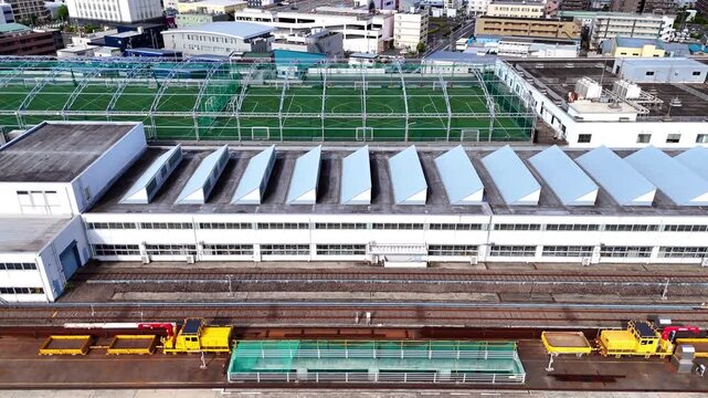 A high-angle aerial perspective looking down at an urban transit depot. The footage reveals the striking geometric pattern of the facility's sawtooth roof, adjacent maintenance tracks, and a rooftop s