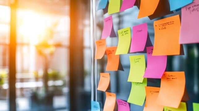 Brightly colored sticky notes, containing handwritten messages, are attached to a transparent glass panel in a contemporary office environment, bathed in warm sunlight