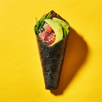 A Japanese temaki sushi cone with tuna, avocado, and sesame seeds on a bright yellow background