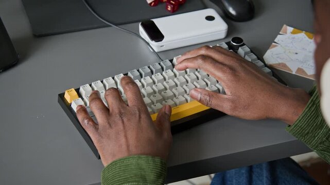 High-angle close-up of hands of unrecognizable African American male software engineer writing prompt for vibe coding agent on keyboard while designing mobile application for tablet