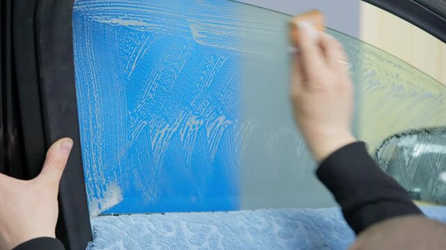 Close-up view of a skilled worker meticulously washing the interior of a car side window with a soapy sponge, preparing the glass surface for tinting film installation in a professional workshop