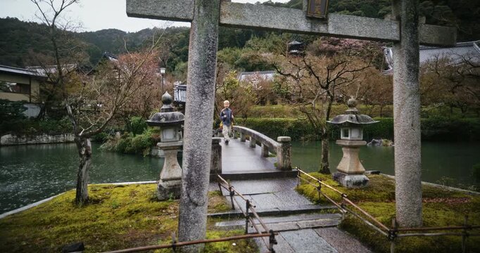 Epic swooping shot around ancient Japanese torii gate and bridge in village as child walks across it in the rain