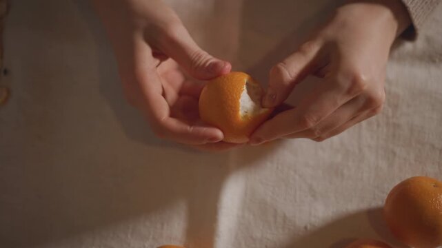 Hands peeling orange revealing white pith and segments on linen table, warm moody light with tactile close framing, deliberate fingers separating rind, sensory cozy moment focused on texture and taste