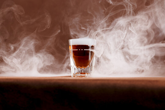Whiskey in a traditional shot glass surrounded by a smokey bar top in a brownish, old western feel to the ambience. Intentionally shot with a warmer off camera lighting to emulate a traditional bar