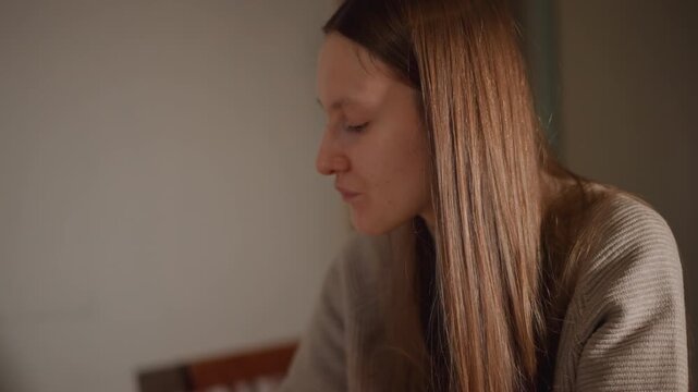 Caucasian woman quiet contemplation at desk soft warm light highlights long straight hair and wool sweater interior corner student reviewing notes writer drafting pages person pausing in thought