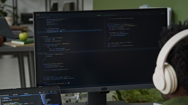Medium over-shoulder shot of busy African American male software developer in headphones writing algorithm on desktop computer while designing mobile app, tablet with website code on desk