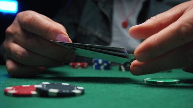 Hands of a male poker player reveal playing cards while placing chips on a green felt table in a dimly lit casino environment