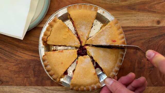 Closeup overhead shot of hands using a tool to cut the final slice from a sugar coated, baked dessert pie &ndash; crisp and crumbly on the outside, with red fruit filling.