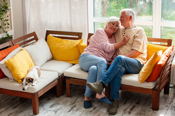Senior woman and senior man sitting on sofa at home with dog. Happy elderly couple hugging in...