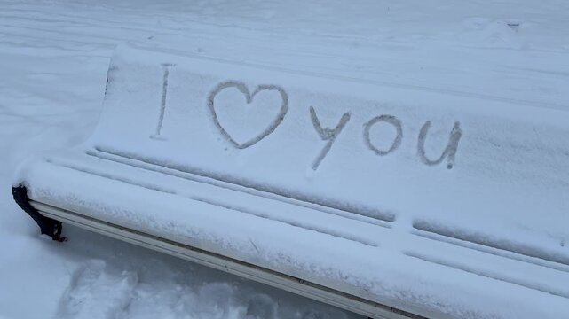 Words I love you drawn with a heart symbol in fresh white snow on a wooden park bench.