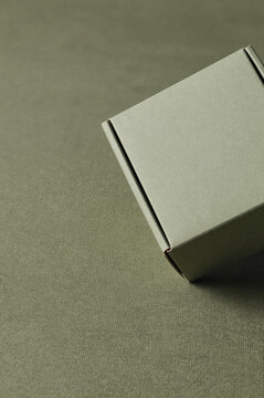 Cardboard box rests on a textured fabric surface in an angled composition. Soft shadows fall across the sage green packaging and grainy textile under diffused daylight.