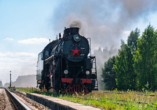 Old black steam locomotive moving along parallel train tracks next to a dense green forest