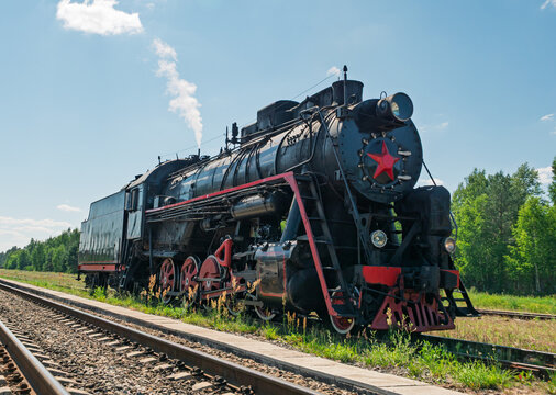 Old black steam locomotive moving along parallel train tracks next to a dense green forest