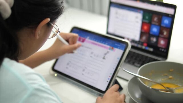 Cinematic over the shoulder shot of a young girl with glasses using a stylus pen on tablet, studying online with laptop at home