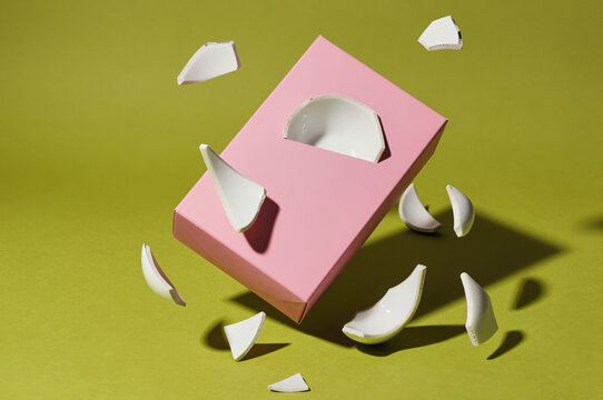 Pink box floats in midair against a lime green backdrop while white ceramic shards revolve around its center in sharp focus under harsh sunlight with deep shadows.