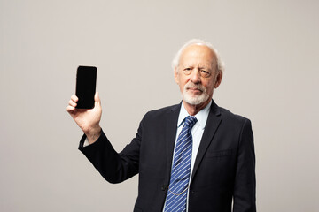 Naklejka premium Senior white man in a suit holding up a smartphone on a plain background