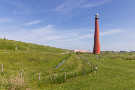 Den Helder, Netherlands. A wide scenic view of the historic red Lange Jaap lighthouse standing against a blue sky, surrounded by green grassy dikes and a walking path.