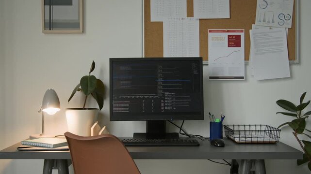 Medium full no people shot of desk of software engineer at startup technology company office, with computer code running on black screen, lamp, pot plant, pin board with project documentation