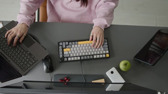 Medium top view of hands of anonymous young female software developer, dressed in pink sweatshirt, typing code on computer, testing script on laptop and tablet while working on multiple devices