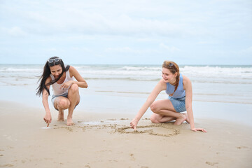Fototapeta premium Young women are squatting on a wet sandy beach, looking down at something they are interacting with in the sand, with the ocean and a cloudy sky in the background
