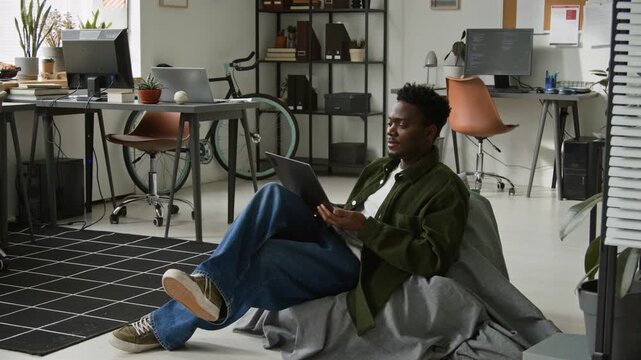 Full shot of cheerful young zoomer African American software engineer sitting down in bean bag chair in office with laptop computer to work with AI vibe coding agent on mobile application project