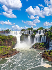 Breathtaking scenic view of the majestic Iguazu Falls with cascading water, lush green cliffs, and a vibrant blue sky with fluffy white clouds © Yuliko