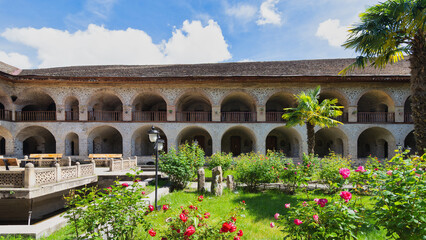 Obraz premium Historic Upper Caravanserai courtyard in Shaki, Azerbaijan, featuring arched stone architecture, vibrant rose gardens, a palm tree, and a sunny blue sky