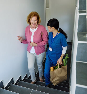 Doctor or nurse caregiver helping senior woman walking up the stairs and carrying groceries at home or nursing home