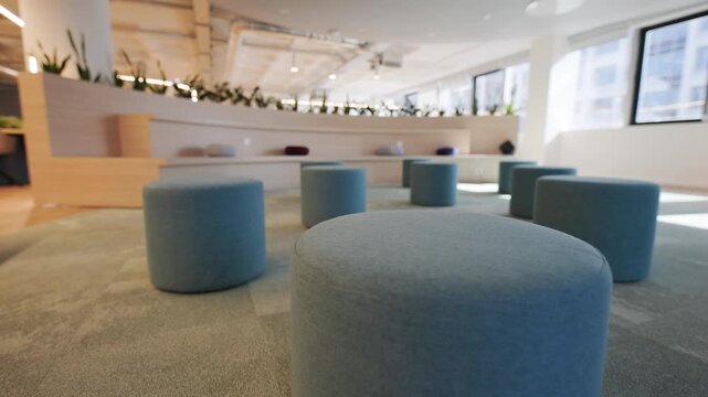 Modern office interior featuring circular blue stools arranged on a soft carpet, with a wooden seating area and greenery in the background creating a contemporary workspace atmosphere