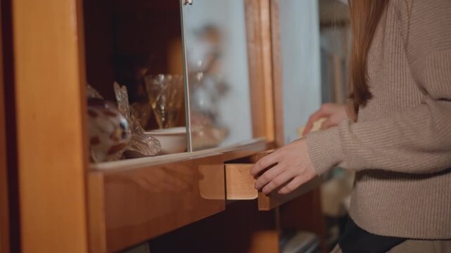 Woman hands opening wooden drawer revealing napkin and glassware long hair and knit sweater, warm amber lighting, vintage cabinet, gentle cleaning motion hostess prepares table, collector inspects