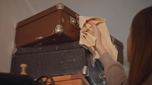 Woman dusting top suitcase on wardrobe shelf with soft cloth, careful strokes on leather corners, focused motion and steady hands near sewing machine and wooden decor, muted warm palette