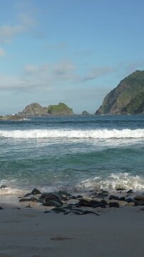 Scenic vertical shot of a pristine tropical coast with turquoise sea water, rolling waves, rocky hills, and a clear blue sky. Papuma Beach, Jember, Indonesia.