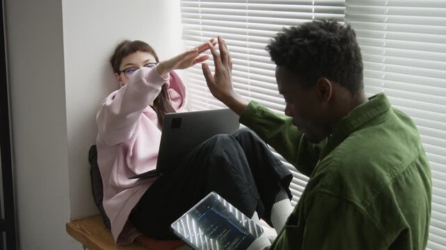 Medium shot of multiethnic mobile app developers testing code on laptop and tablet, Caucasian woman cheering and giving high fives to African American man at successful prototype implementation