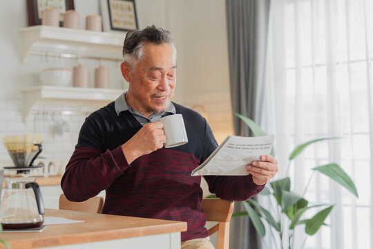Happy senior Asian man using laptop and holding coffee cup inside home kitchen concept for working retirement independent lifestyle digital literacy financial freedom smart elderly citizen