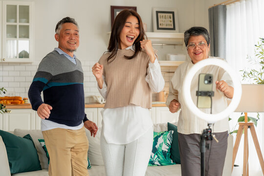 Happy Asian multi-generation family making vlog video live streaming online with smart phone and ring light inside home kitchen concept for social media influencer digital content creator technology m