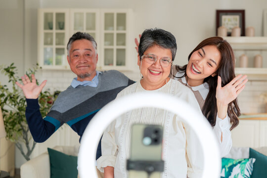 Happy Asian multi-generation family making vlog video live streaming online with smart phone and ring light inside home kitchen concept for social media influencer digital content creator technology m