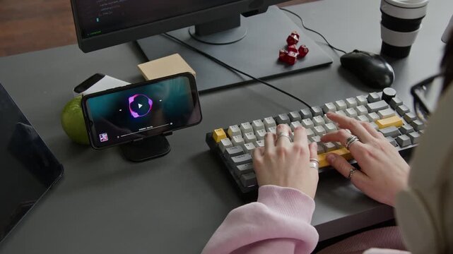 Over-shoulder shot of woman in wireless headphones listening to music on smartphone, while typing on keyboard and writing computer code, suggesting inspiration and motivation for work