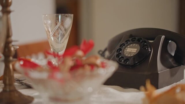 Vintage rotary phone on table with crystal glass and candy bowl, warm evening light, idle handset resting beside lace tablecloth, timeless domestic stilllife evoking memory and silent waiting
