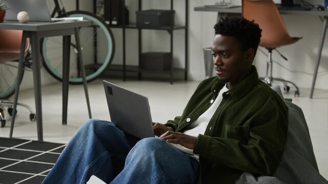 Medium full shot of young gen Z African American programmer writing computer code on laptop with advantage from vibe coding assistant, while sitting comfortably in bean bag chair in office