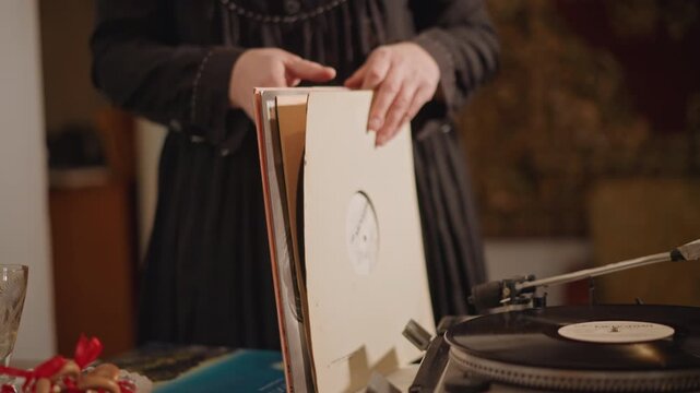 Woman selecting vinyl from stack beside turntable, careful hands flip cardboard sleeves and inspect labels, table with snacks and vintage decor creates cozy collector atmosphere and tactile nostalgia.