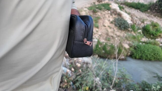 Black PU leather men&rsquo;s clutch with wristlet strap and zippered pocket outdoors in nature.
