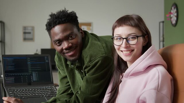 Medium close-up portrait of African American male and Caucasian female programmers discussing project by computer with code, looking at camera, two diverse professionals collaborating in office