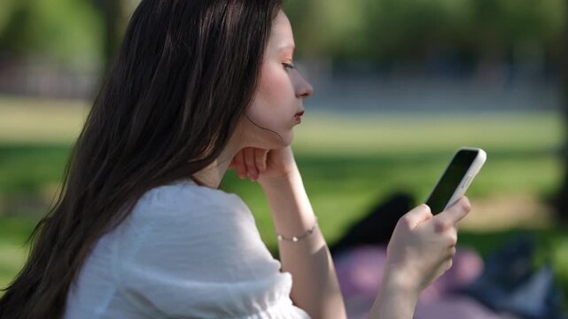 Profile view of a thoughtful young woman with long hair sitting outdoors on a sunny day. She is holding and looking at her cellphone