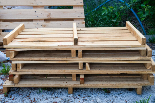 A stack of clean, unpainted wooden shipping pallets rests on a gravel surface outdoors with blurred green foliage in the background.