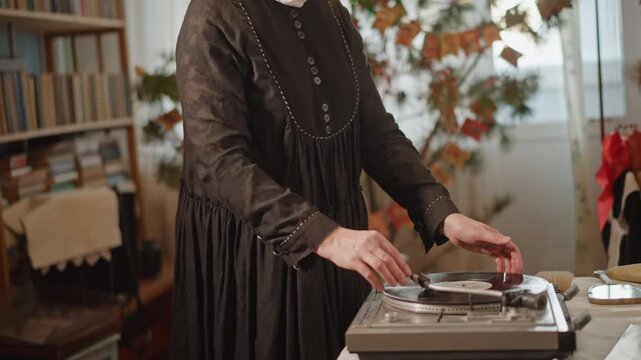 Woman spinning record on turntable, elder archivist in headscarf and glasses working beside bookshelf in cozy home library, focused gentle gestures placing needle, soft natural light and warm